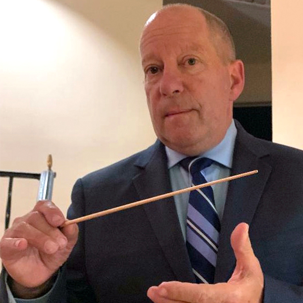 man in dark suite and blue shirt and tie holding a conductor's baton