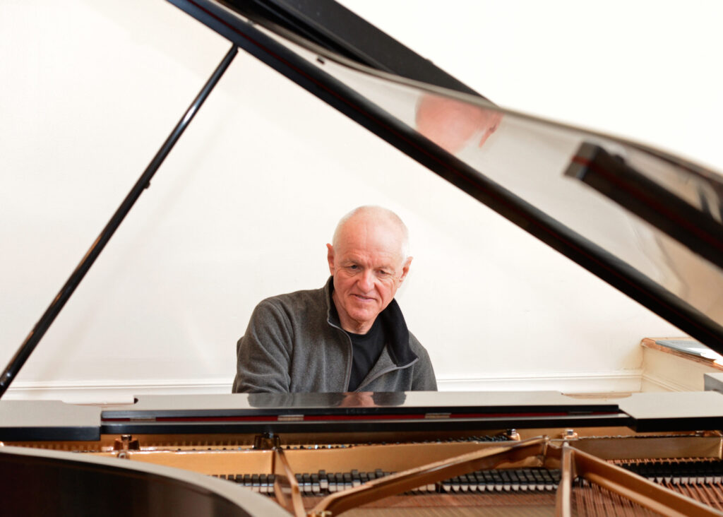 Man sitting at piano viewing man through open lid of the piano
