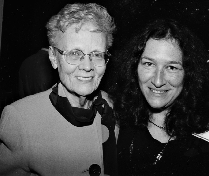 Black and white photo of two women, one with white hair and glasses and one with dark curly hair smiling.