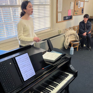 Girl singing next to a piano while a boy observes