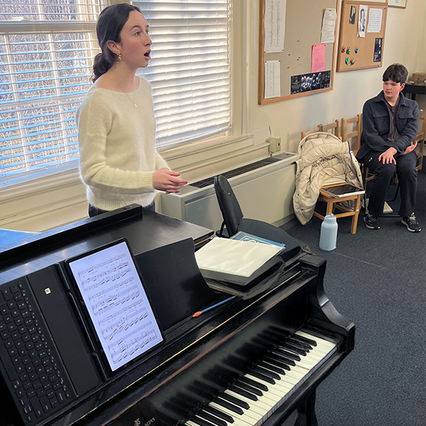 Girl singing next to a piano while a boy observes